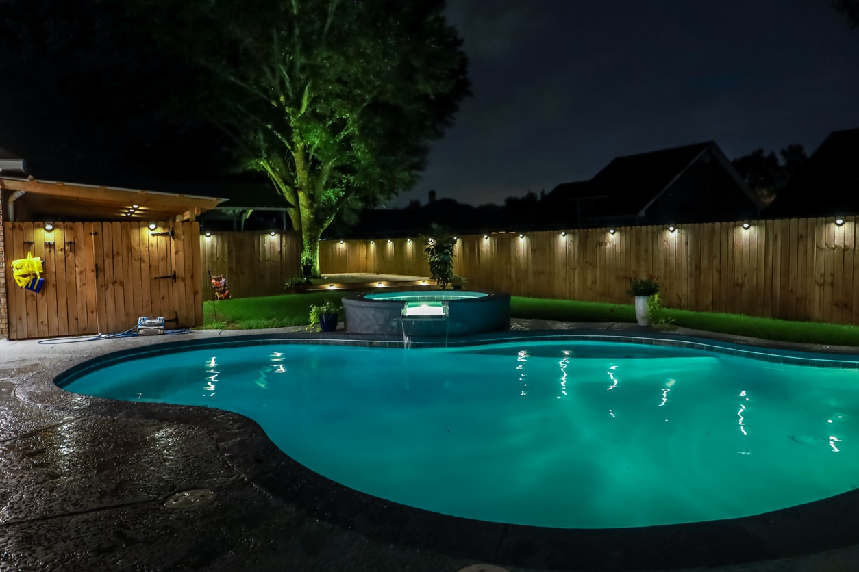 A lit backyard pool at nighttime surrounded by several lights.